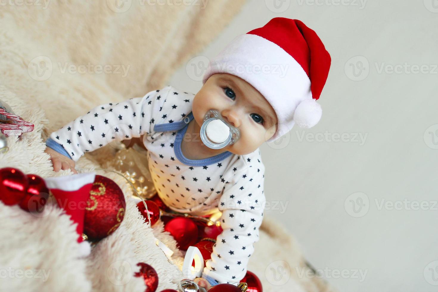 linda niñita con sombrero rojo de santa claus con chupete está jugando con adornos navideños rojos y blancos y luces navideñas en una manta beige. foto
