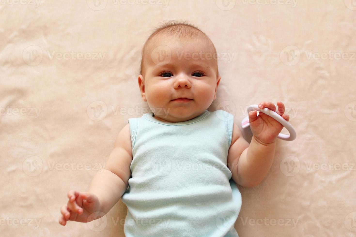 Smiling three month baby in blue bodysuit is lying on a beige blanket and looking at the camera. photo