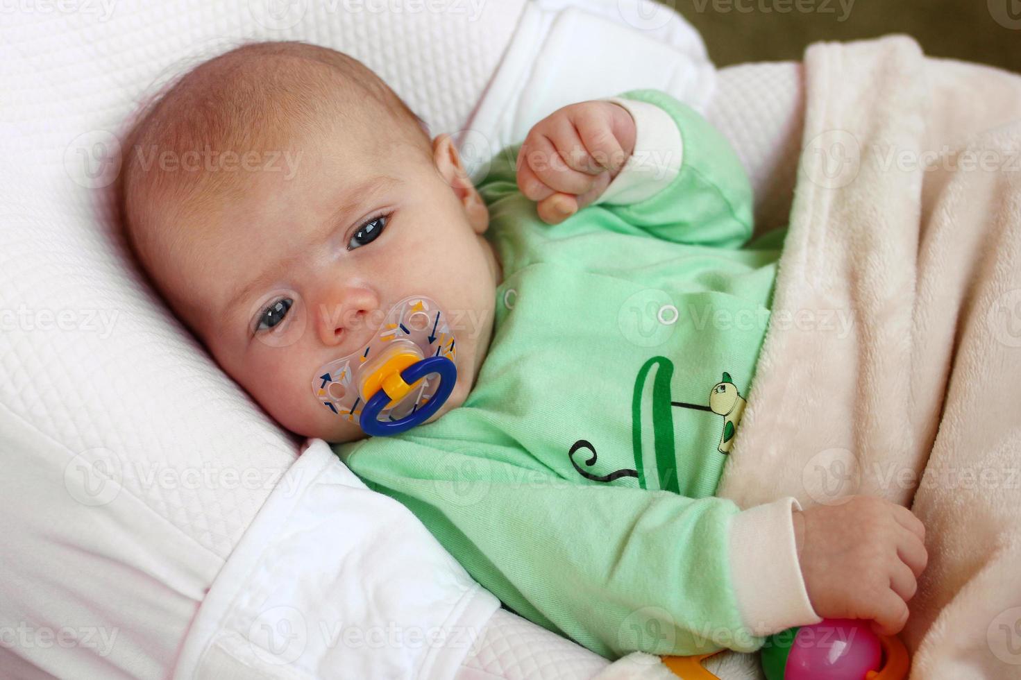 Cute infant little girl with pacifier is lying in her bed under beige plaid. photo