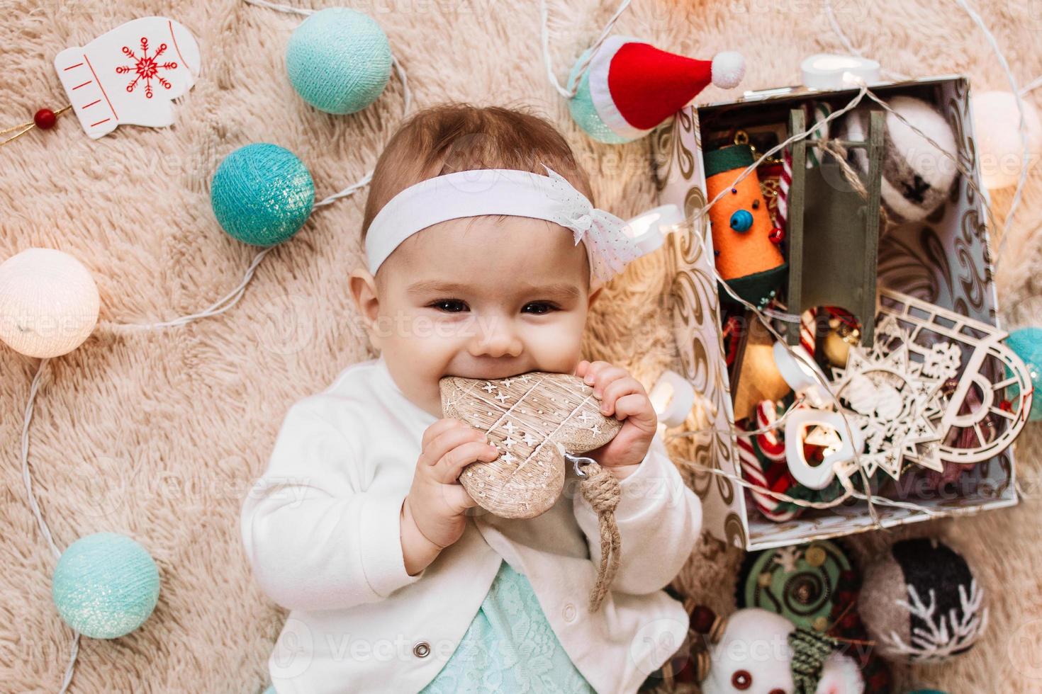 Cute smiling little girl in blue and white dress is playing with wooden heart on a beige plaid with Christmas decorations and lights, top view. photo