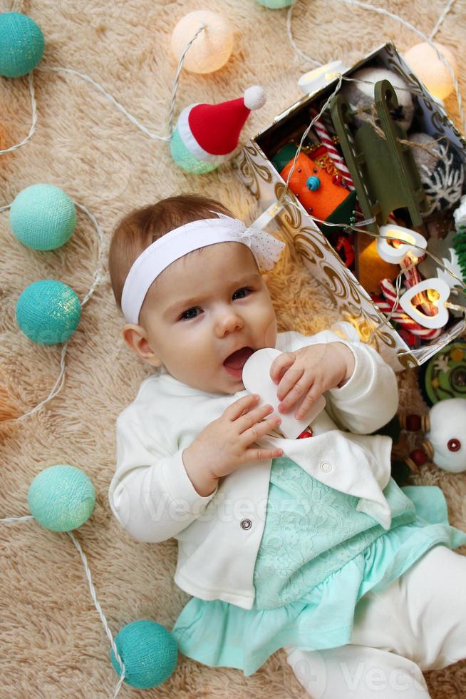 Adorable little girl in blue and white dress is playing with wooden toy on a beige plaid with Christmas decorations and lights, top view. photo