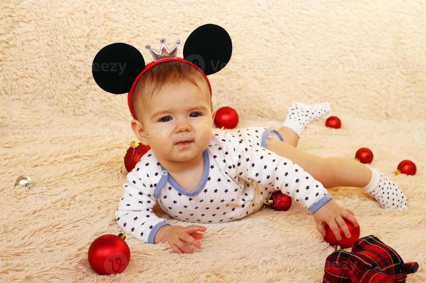 una linda niñita con orejas de ratón está acostada sobre una manta beige y jugando con adornos navideños rojos. foto