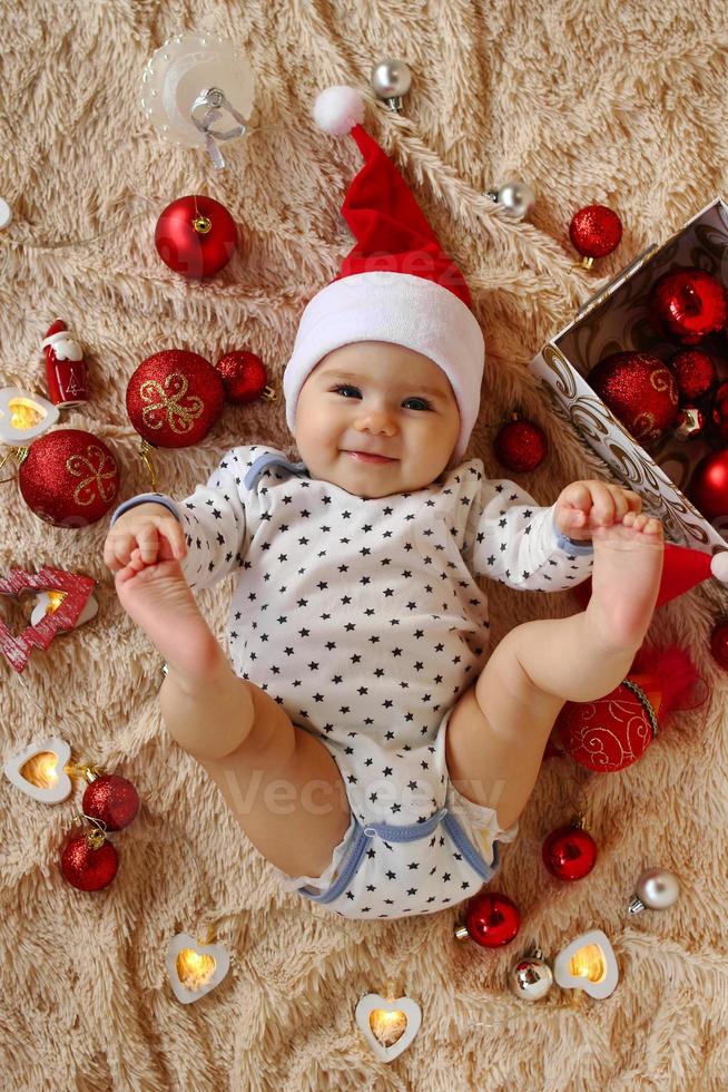 Adorable smiling little girl in Santa Claus red hat is laying on a beige plaid with red and white Christmas decorations and Christmas lights and holding her toes in hands, top view. photo