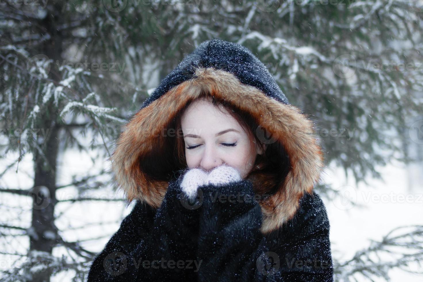 retrato de una joven hermosa mujer con ojos azules en abrigo de piel sintética negra con capucha roja en el fondo del parque nevado de invierno. foto