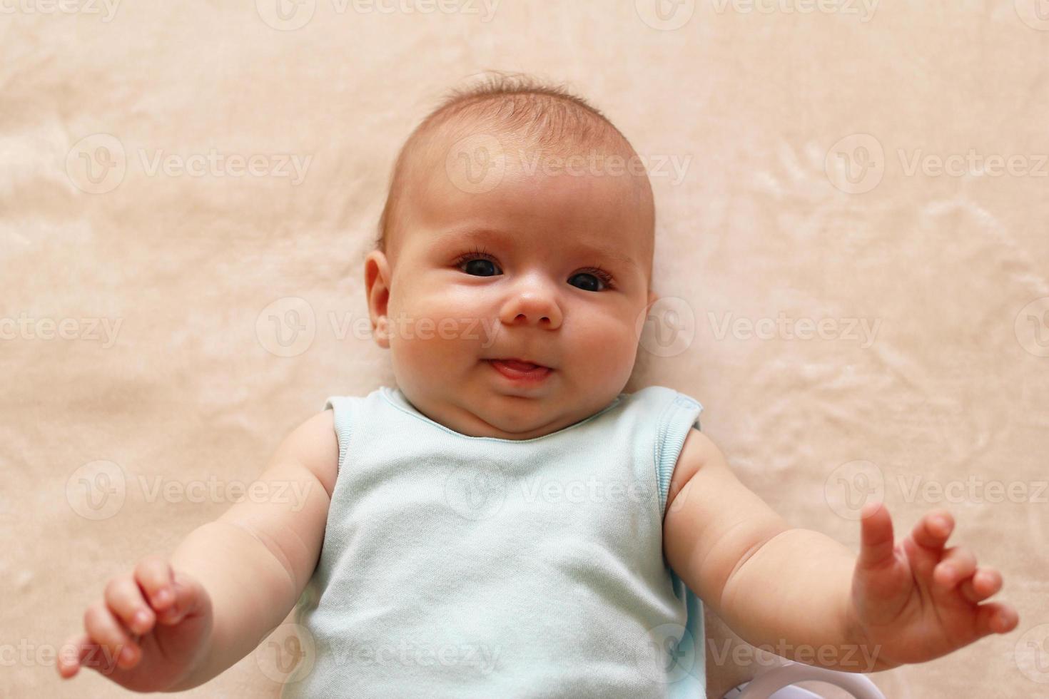 Portrait of smiling three month baby in blue bodysuit is lying on a beige blanket and looking at the camera. photo