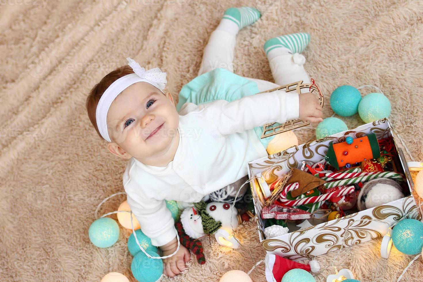 Beautiful smiling little girl in blue and white dress is lying on a beige plaid, playing with Christmas decorations and looking into the camera. photo