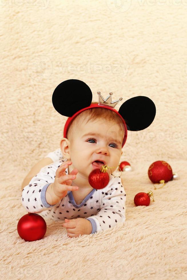 Adorable little girl with mouse ears is lying on a beige plaid and playing with red shiny Christmas balls. photo