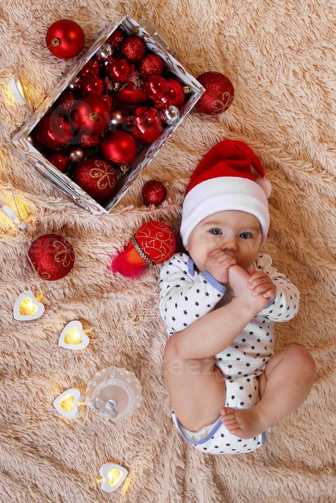 Top view on a little girl in Santa Claus red hat is laying on a beige plaid with red and white Christmas decorations and Christmas lights and holding her foot in her mouth. photo