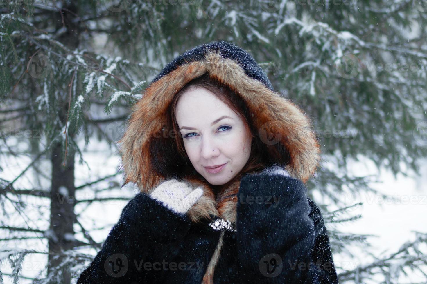 Young smiling beautiful woman with blue eyes in black faux fur coat with red hood on background of winter snowy park. photo