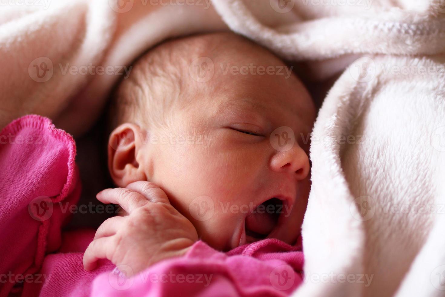 Cute newborn baby in pink baud with an open mouth on her bed under beige blanket. photo