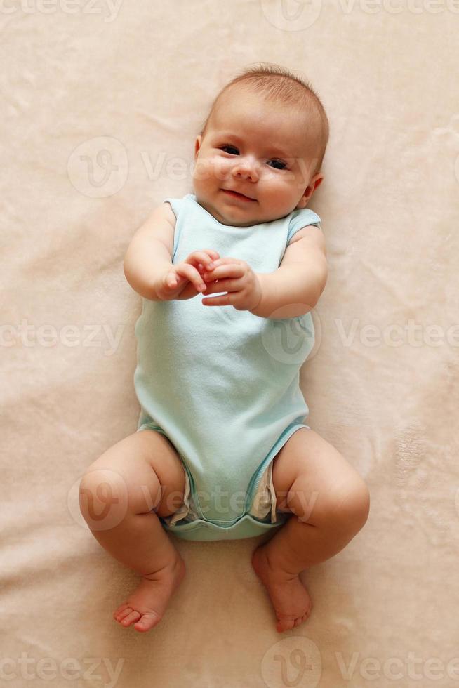 lindo bebé sonriente de tres meses en traje azul está acostado sobre una manta beige y mirando a la cámara. foto