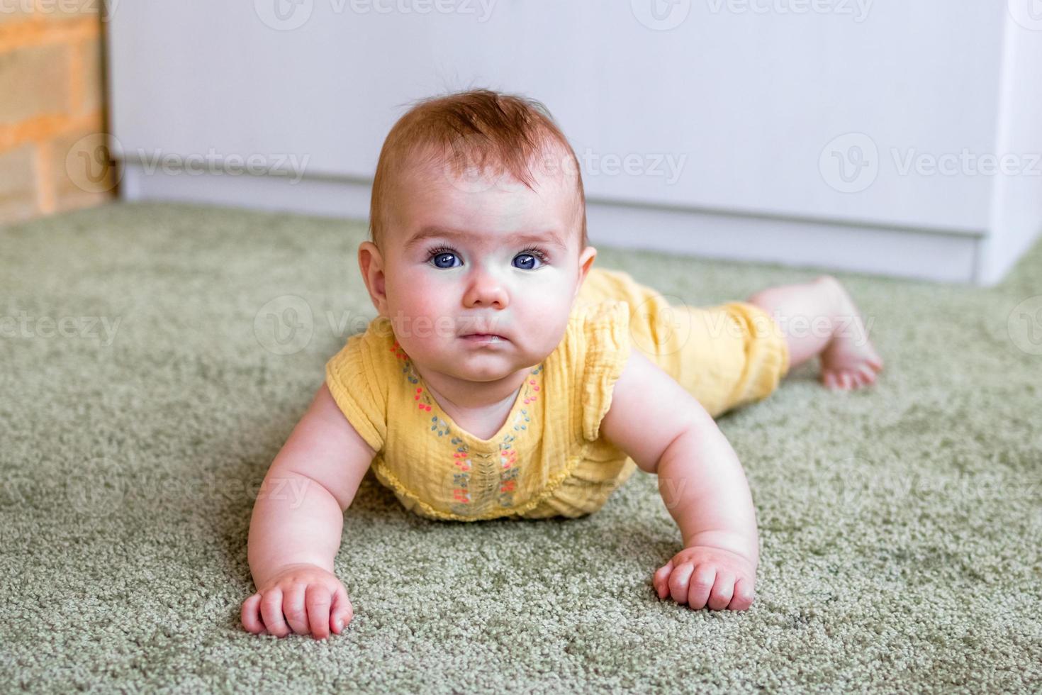 Portrait of little Caucasian baby girl in yellow dress. Child trying to crawl on a floor. photo