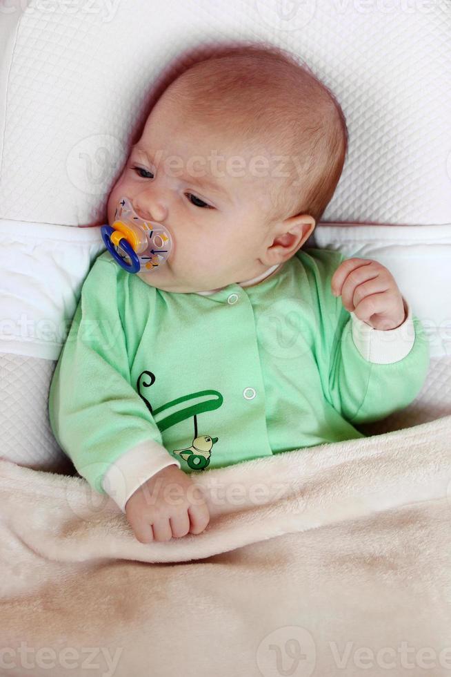 Cute infant little girl with pacifier is lying in her bed under beige plaid. photo