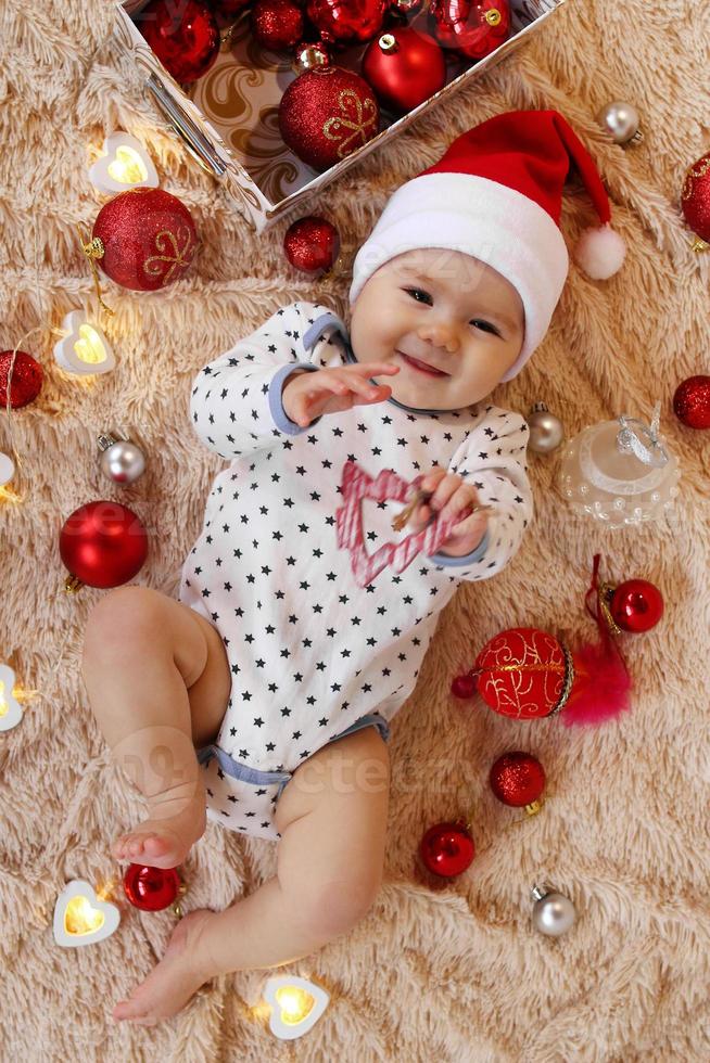 Cute smiling little girl in Santa Claus red hat is playing with wooden toy on a beige plaid with red and white Christmas decorations and Christmas lights, top view. photo