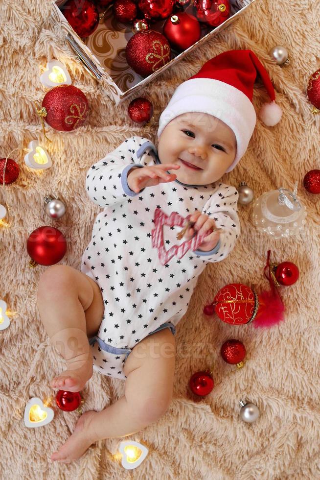 linda niña sonriente con sombrero rojo de santa claus está jugando con un juguete de madera en una manta beige con adornos navideños rojos y blancos y luces navideñas, vista superior. foto