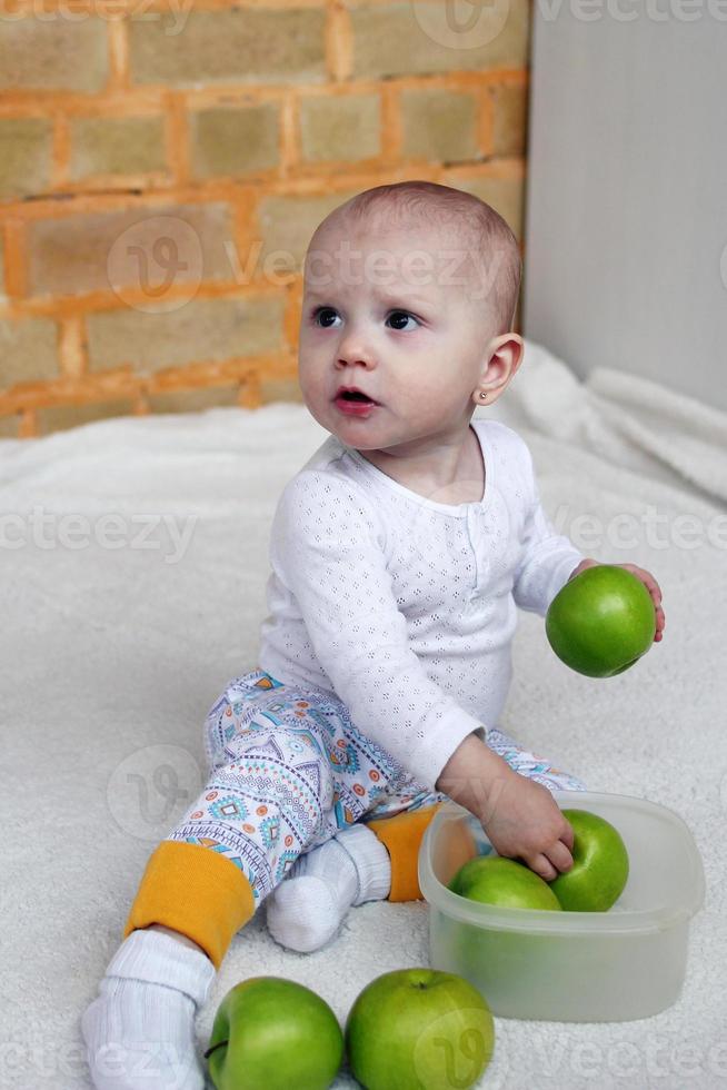 la niña está sentada en la sala de estar sobre una manta blanca con manzanas verdes en un recipiente de plástico. foto