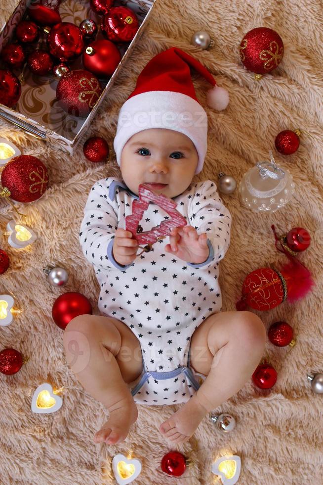 Portrait of cute smiling little girl in Santa Claus red hat is playing with wooden toy on a beige plaid with red and white Christmas decorations and Christmas lights, top view. photo