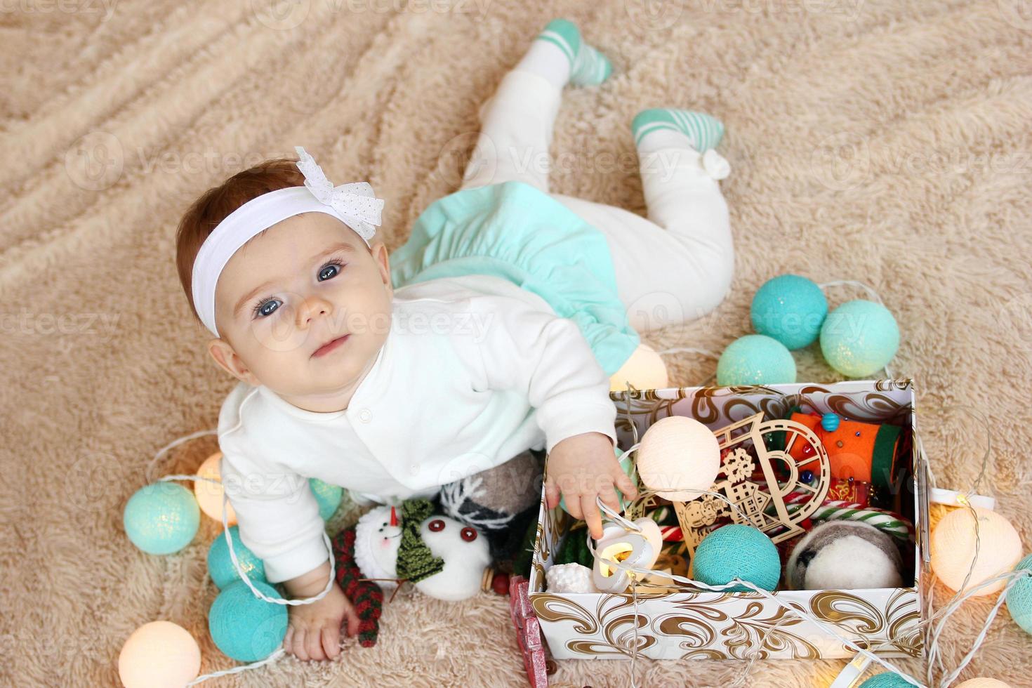 Adorable little girl in blue and white dress is lying on a beige plaid, playing with Christmas decorations and looking into the camera. photo