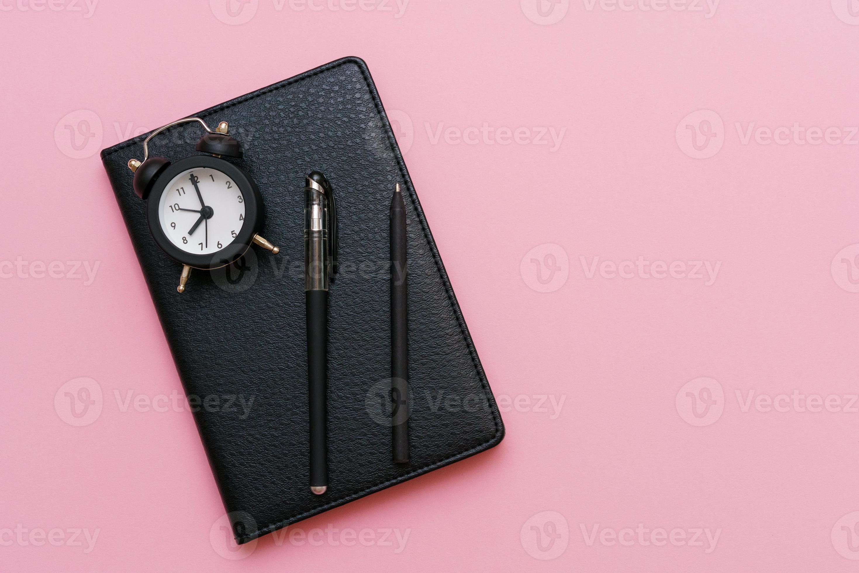 Black notepad with a pen and an alarm clock on a pink background. Using 13396495 Stock Photo at