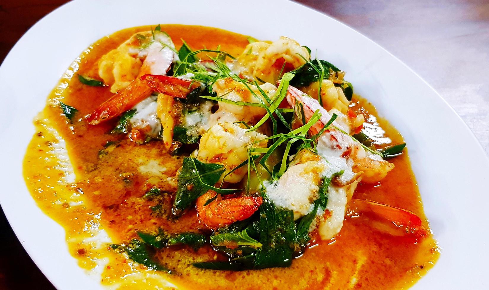 Close up Shrimp on dry red curry with sliced red chili, kaffir lime leaves, basil and herb ingredient on white dish or plate at restaurant. Thai Spicy food photo