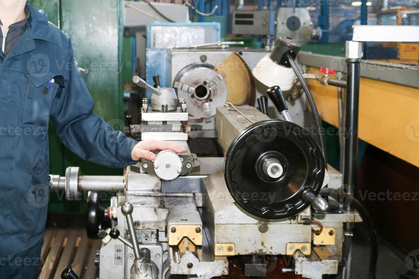 A man working in a robe, overalls stands next to an industrial lathe for cutting, turning knives from metals, wood and other materials, turning, making details and spare parts at the factory photo
