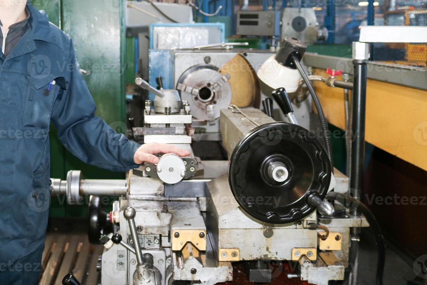 un hombre que trabaja con una túnica, overol se encuentra junto a un torno industrial para cortar, tornear cuchillos de metales, madera y otros materiales, tornear, hacer detalles y repuestos en la fábrica foto