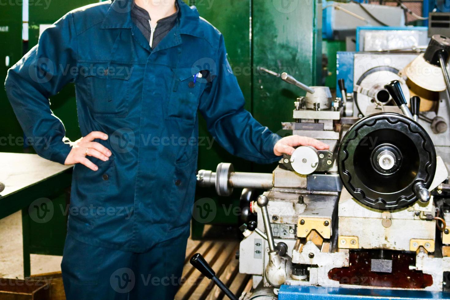A man working in a robe, overalls stands next to an industrial lathe for cutting, turning knives from metals, wood and other materials, turning, making details and spare parts at the factory photo