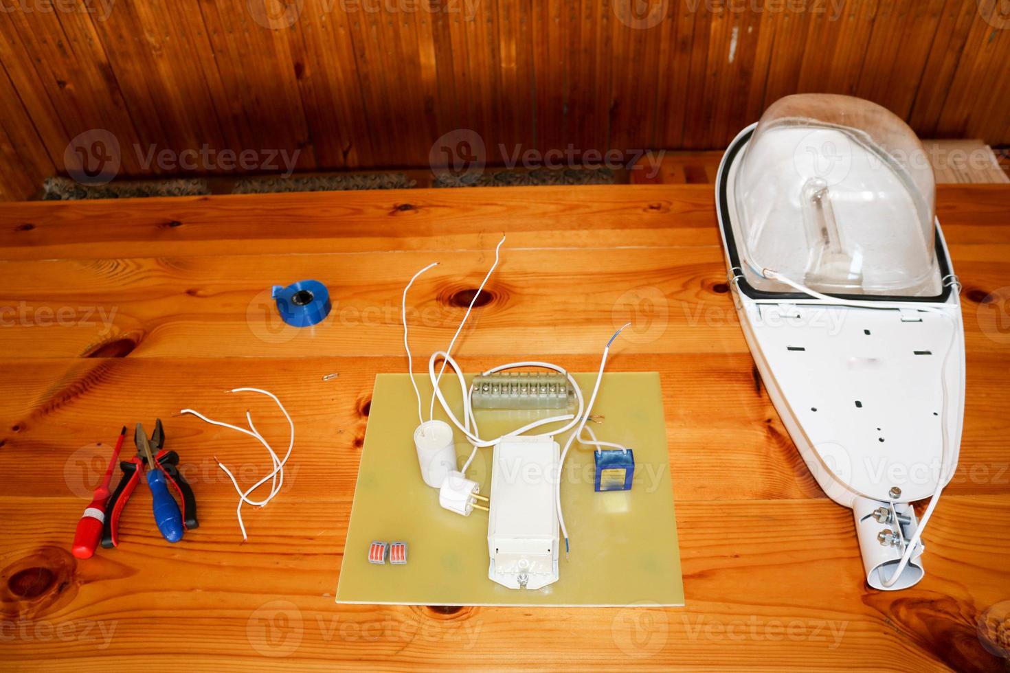 A large white disassembled street lamp with an electrical circuit with wires and spare parts, installation equipment, pliers, blue electrical tape, screwdrivers on the table photo