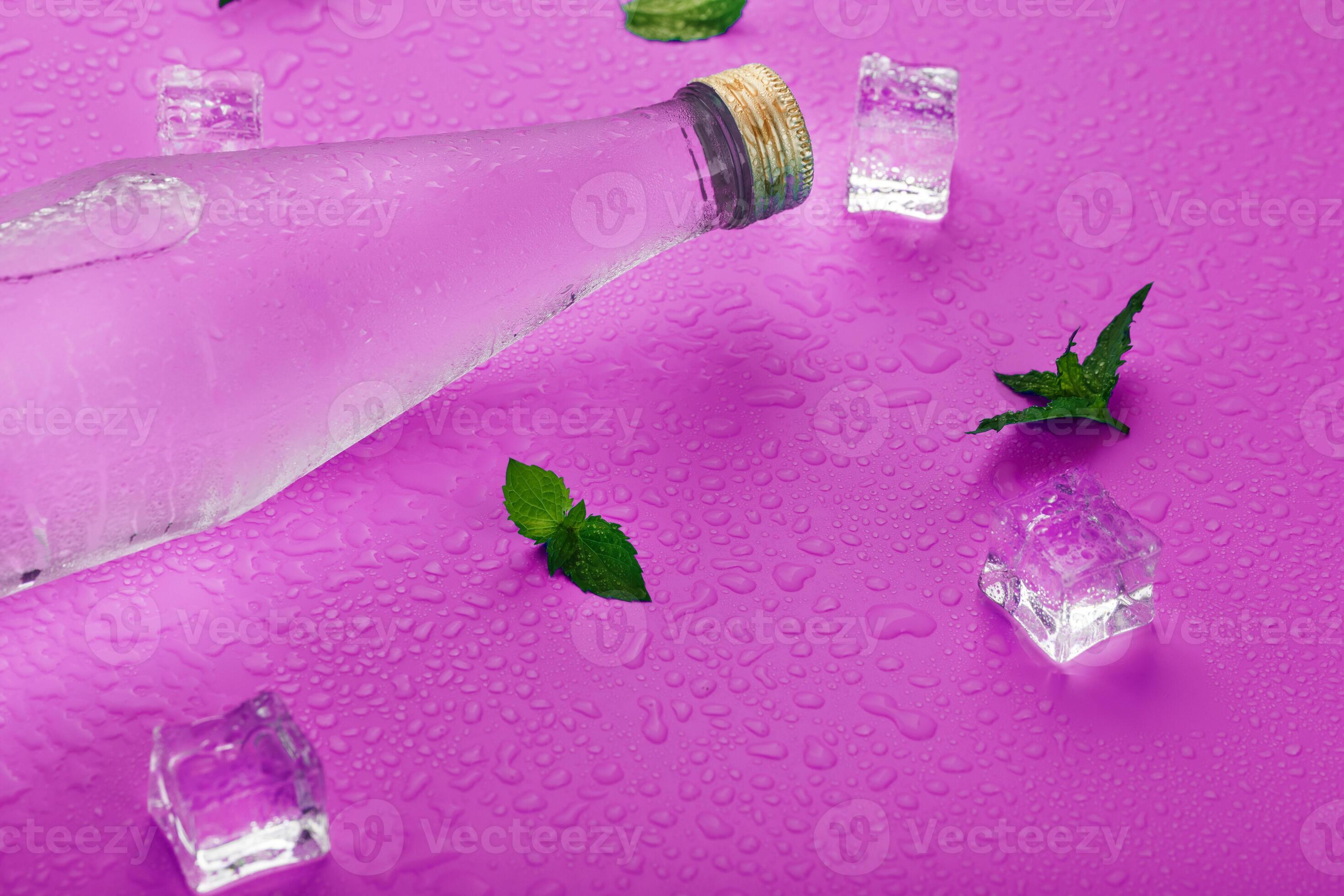 A bottle with an ice drink in drops of condensation, ice cubes and mint leaves on a pink