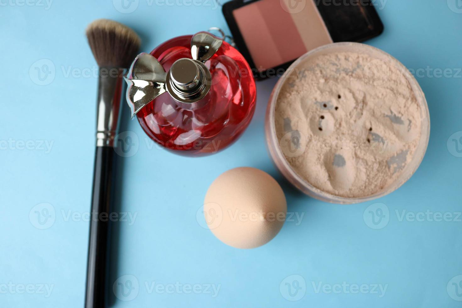 Beauty box, crumbly matte mineral powder and blush with a beauty blender with a brush and red glass bottle of perfume on a blue background. Flat lay. Top view photo