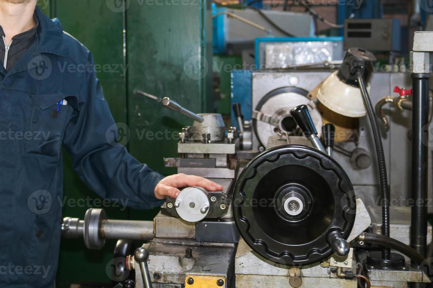 A man working in a robe, overalls stands next to an industrial lathe for cutting, turning knives from metals, wood and other materials, turning, making details and spare parts at the factory photo