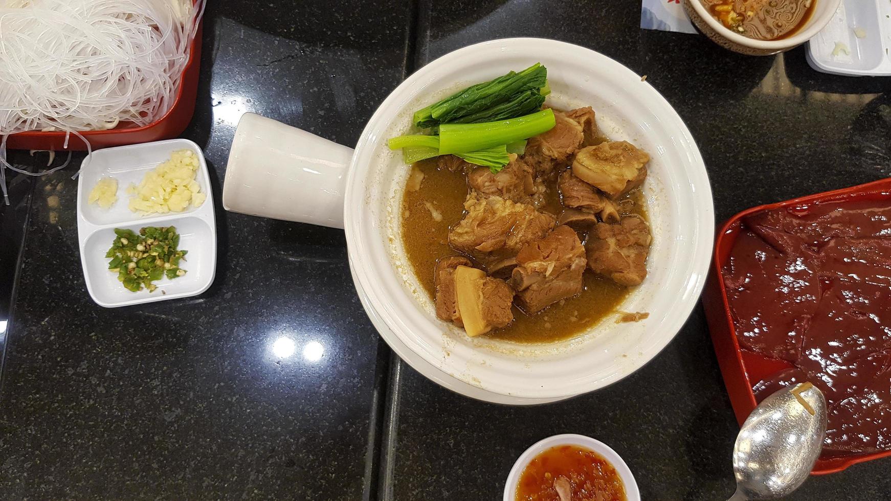 Top view of Braised pork ribs with kale in hot bowl dish and fresh liver in red tray and side dish with sliced green chili and garlic on black dining table. Rice noodle with spicy or sour sauce. photo