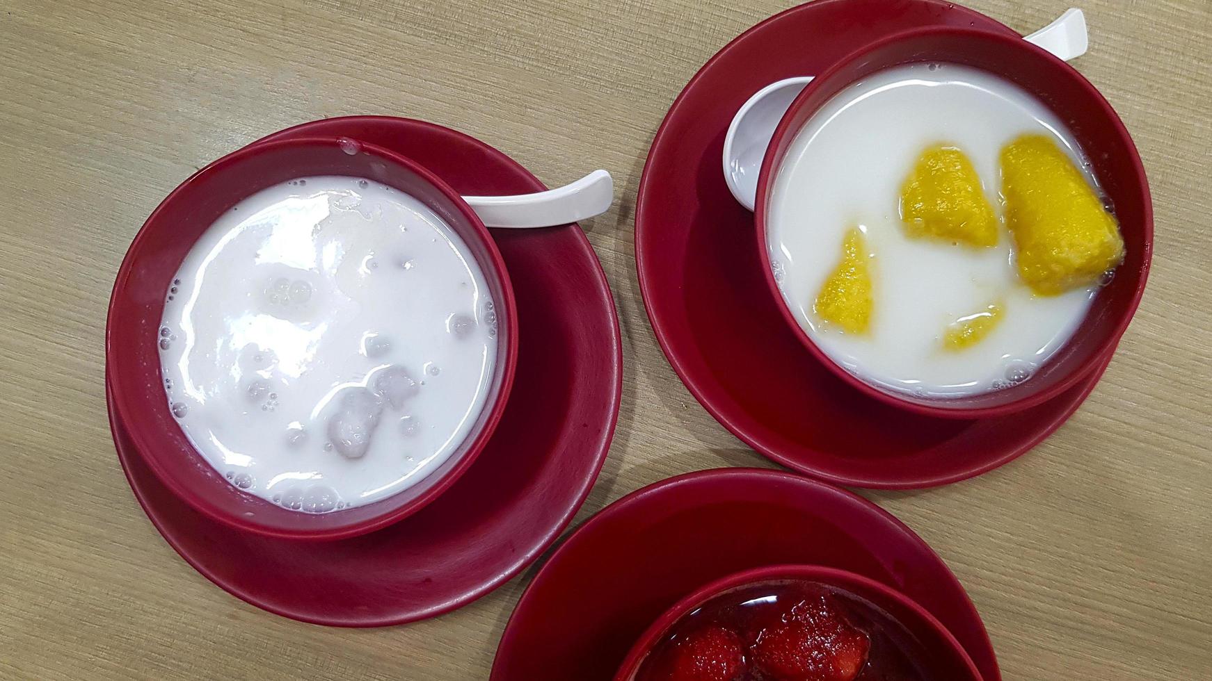 Top view of Thai dessert with strawberry in syrup, banana in coconut milk and taro Dumplings in coconut cream in red bowl and mobile phone or telephone on wooden table or background. Sweet Asian food. photo