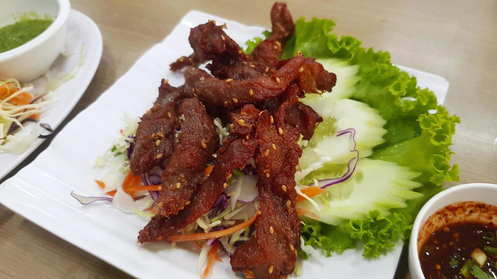 cerdo seco frito con rodajas de pepino, repollo verde en un plato o plato blanco y salsa picante. comida tailandesa o asiática foto