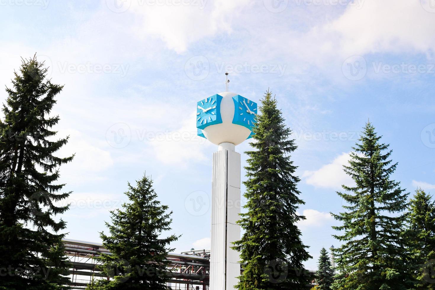 reloj inusual sobre un fondo de un cielo blanco-azul. un reloj en forma de molécula, un átomo. sobre la producción química, una columna de vigilancia, una torre de reloj en un bosque de abetos. telón de fondo de tuberías químicas. foto