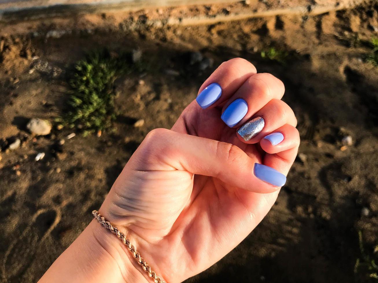 hand with blue manicure on sand background. the ring finger is painted in silvery sequins. a new color of nails for a girl with a gold bracelet. in the background - the construction of a new district photo