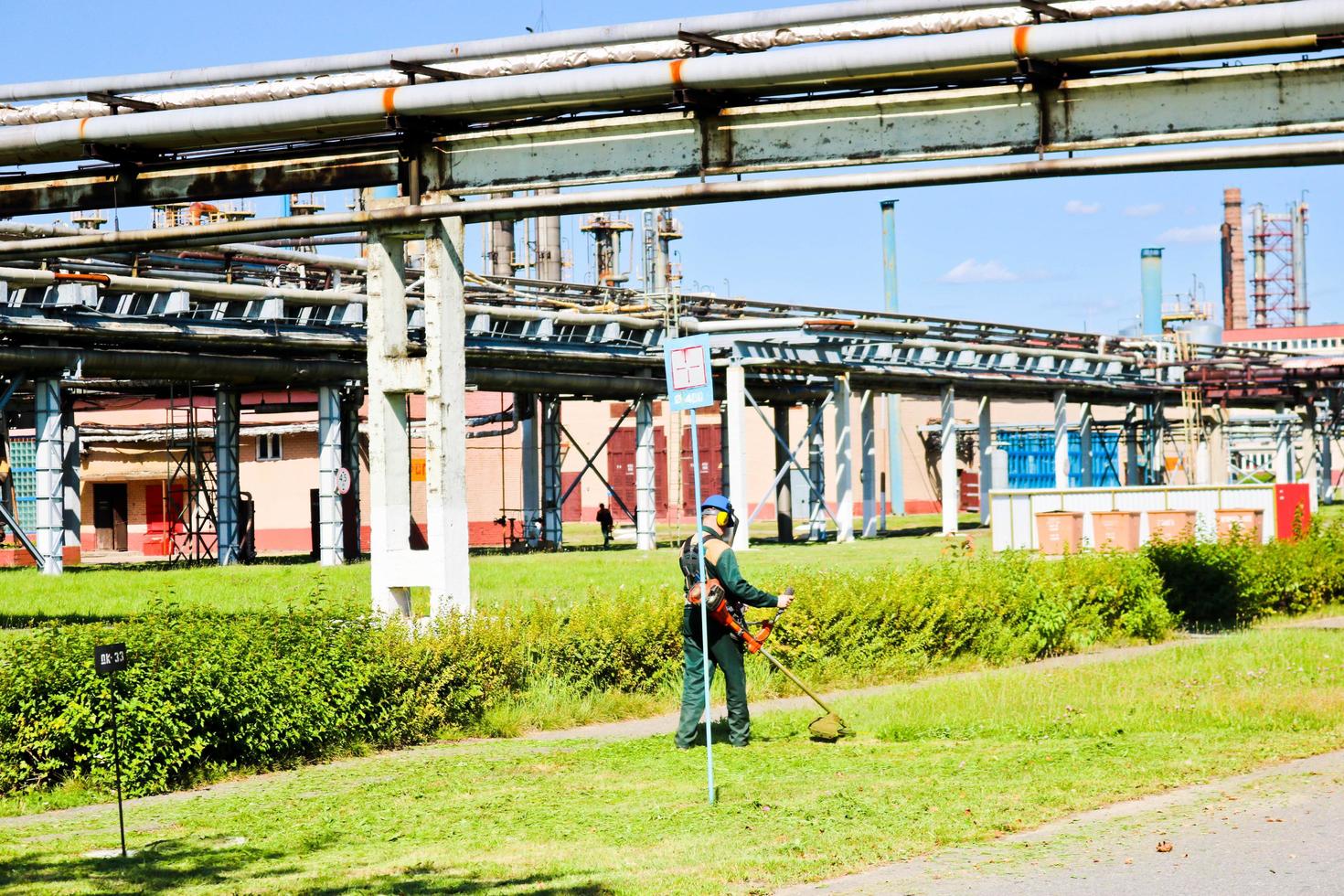 The worker in a protective suit and mask uses a special device to mow the grass on a sunny summer day. Lawn mower in the hands of a man. Against the backdrop of pipes on chemical production photo