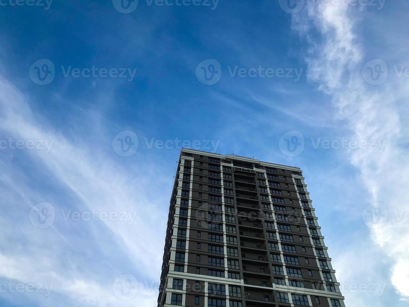 a tall, multi-storey glass building in a white and brown city. the house has balconies, stylish decoration. ergonomic arrangement. against the backdrop of a sunny blue sunny sky photo