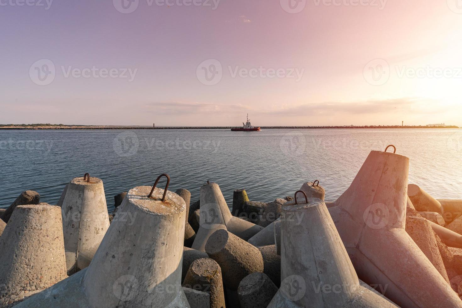 Ship-tugboat goes in high seas to tow cargo ship to port. Beautiful sunset over the pier. Tetrapod breakwaters in harbor. photo