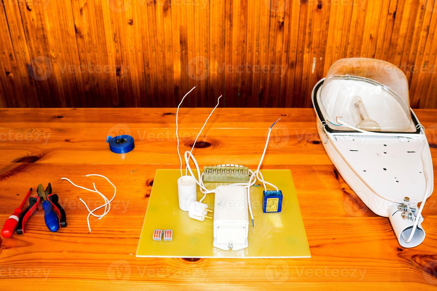 A large white disassembled street lamp with an electrical circuit with wires and spare parts, installation equipment, pliers, blue electrical tape, screwdrivers on the table photo