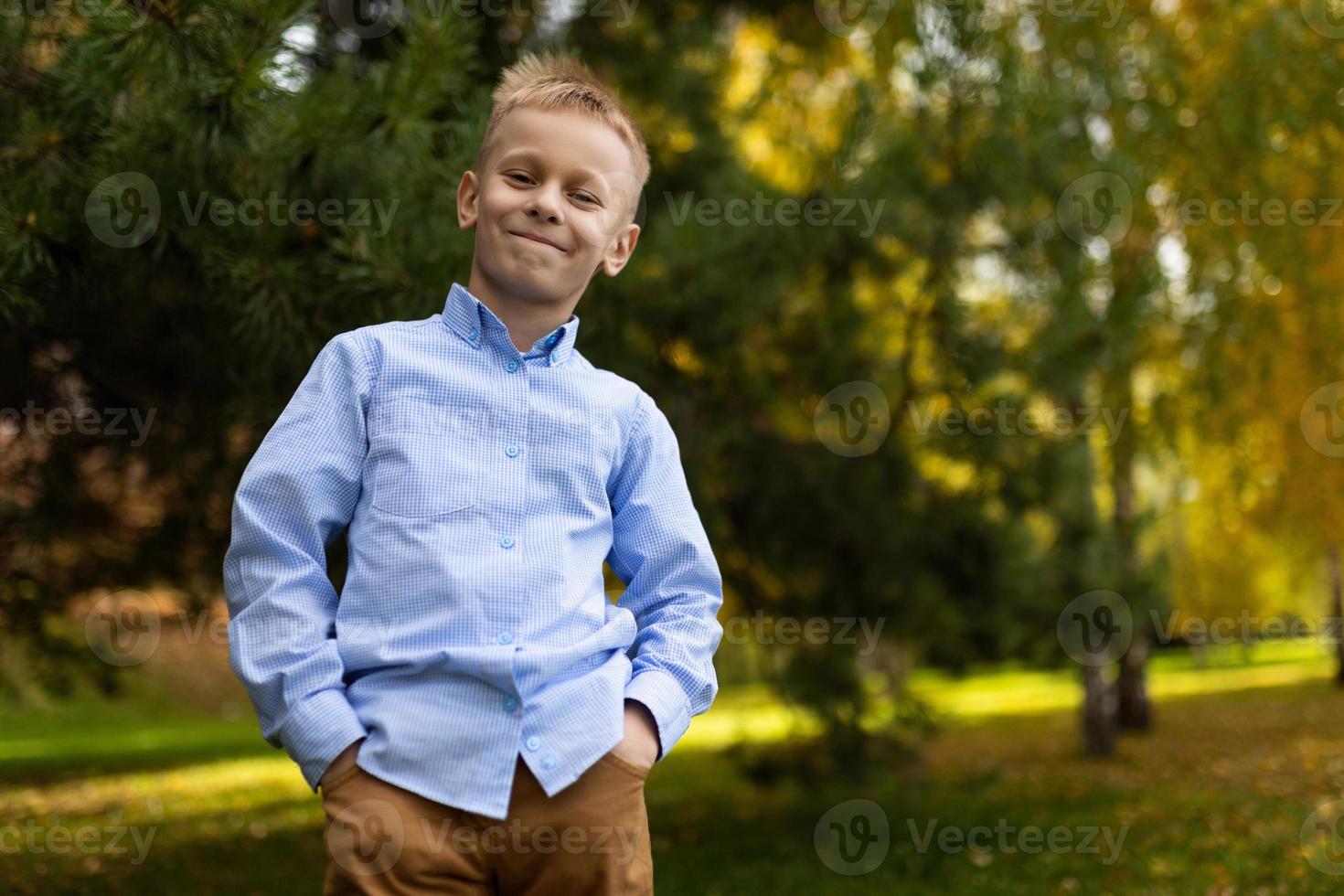 portrait of a cheerful boy in a blue shirt standing with his hands in his pockets against the backdrop of an autumn forest photo