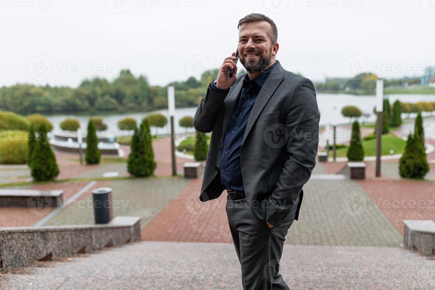 exitoso hombre de negocios satisfecho habla con una sonrisa en un teléfono móvil bien en un traje de negocios en el fondo de un parque de la ciudad foto