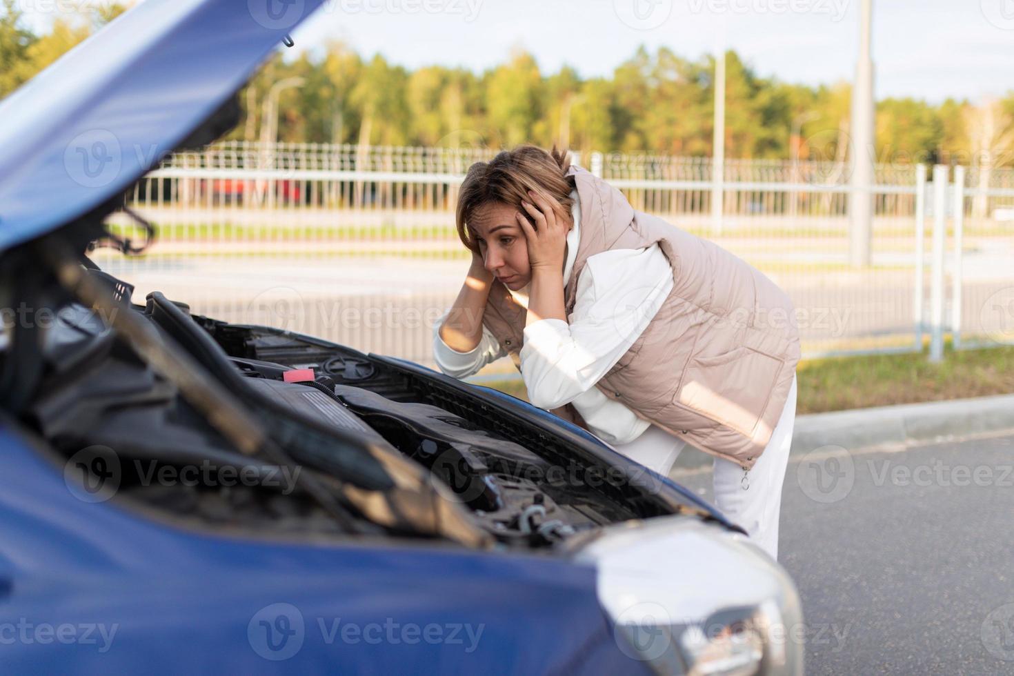 una mujer conductora desconcertada, llevándose las manos a la cabeza, mira el motor de un camión roto foto