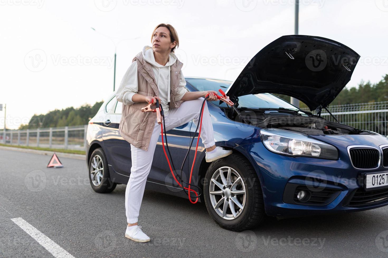 a woman on the road next to a car whose battery is dead and is looking for a person from whom you can recharge the car battery photo