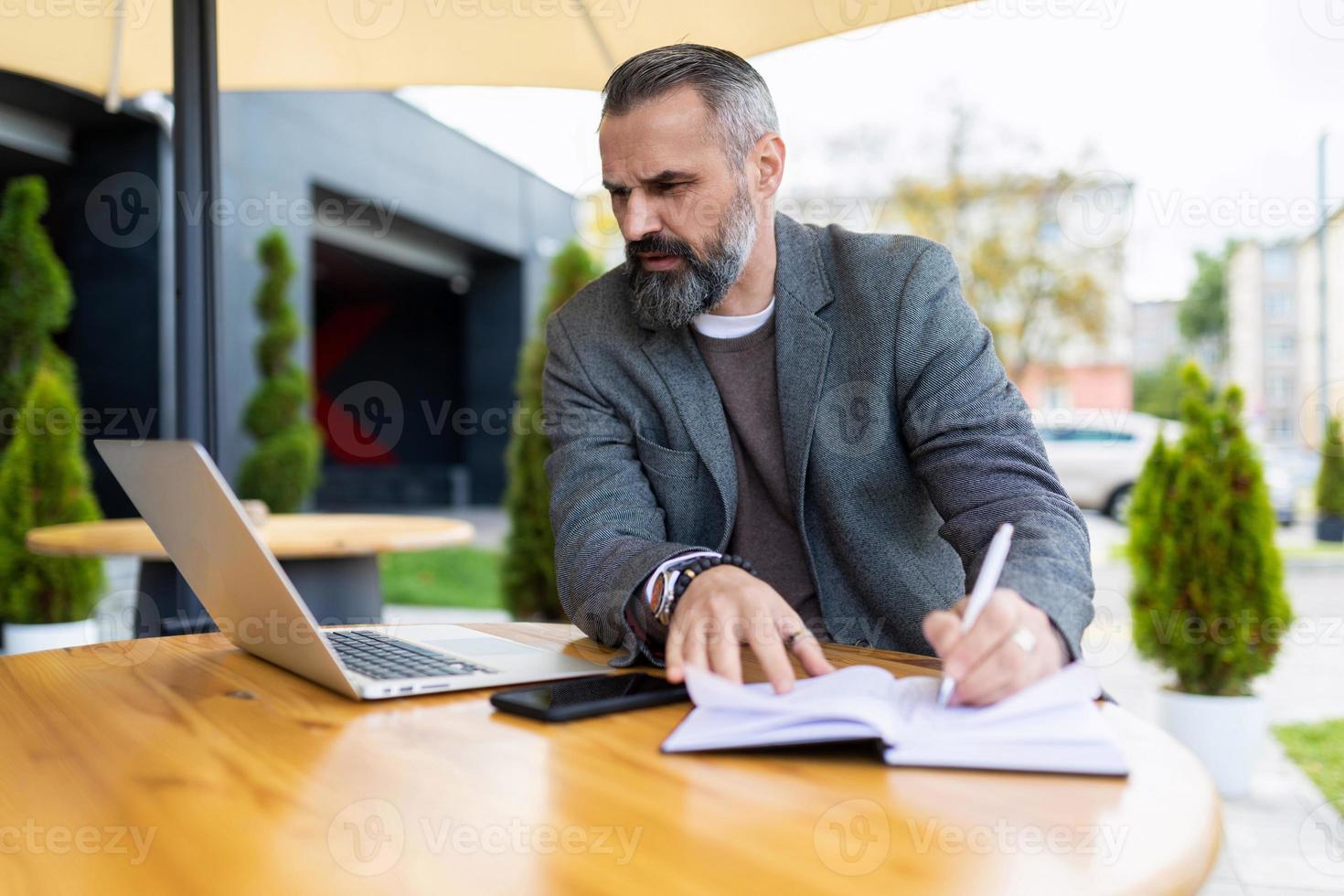 hombre de negocios adulto que trabaja en una computadora portátil en línea en un café de la ciudad al lado de un edificio de oficinas, concepto de trabajo independiente y remoto foto