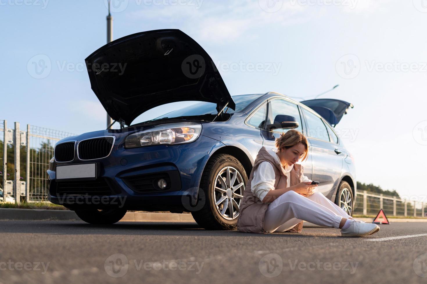 frustrated woman sitting on the road near the broken down car with the hood up, car insurance concept photo