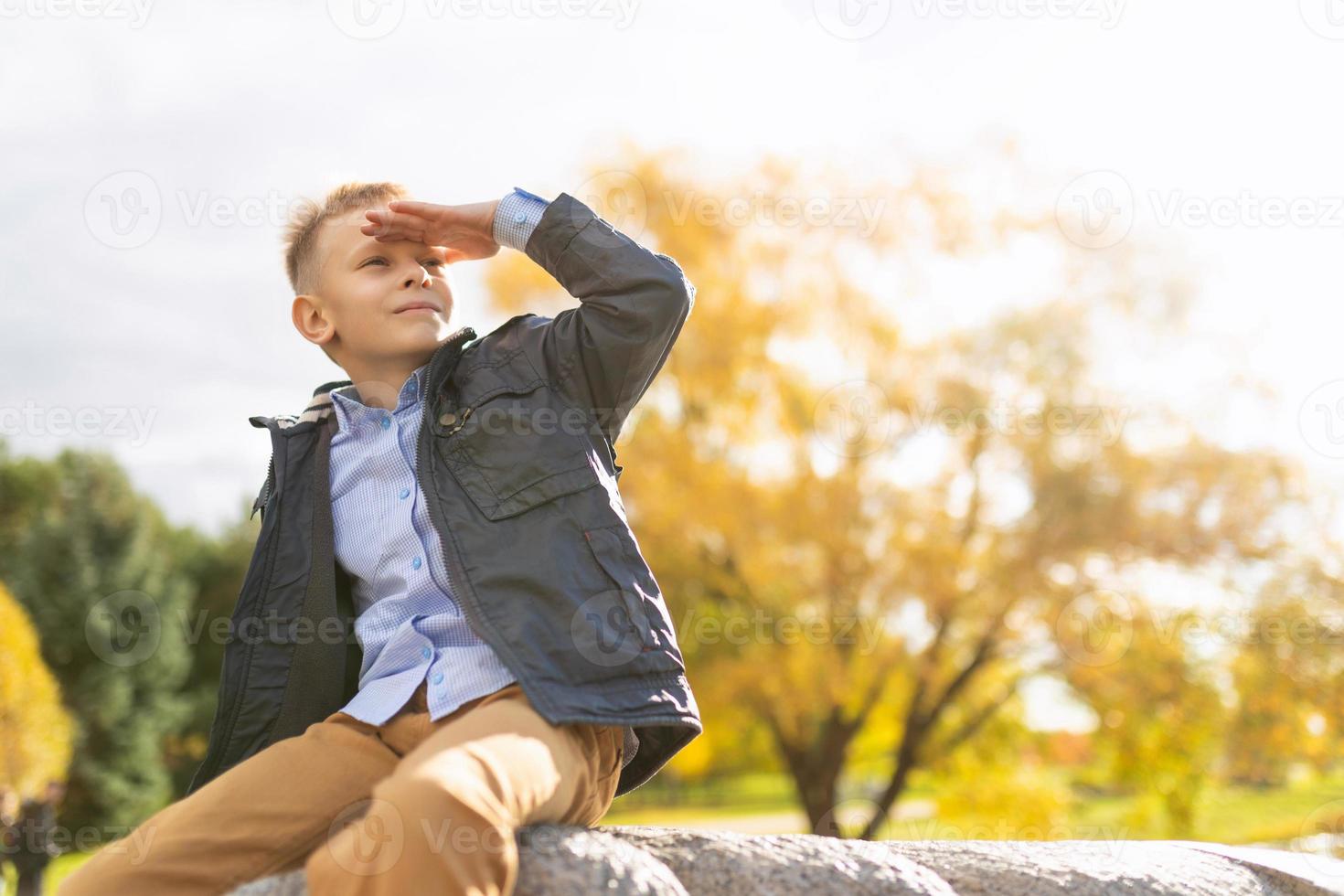 Cheerful mischievous boy in the autumn park looks into the distance with his hand to his forehead photo