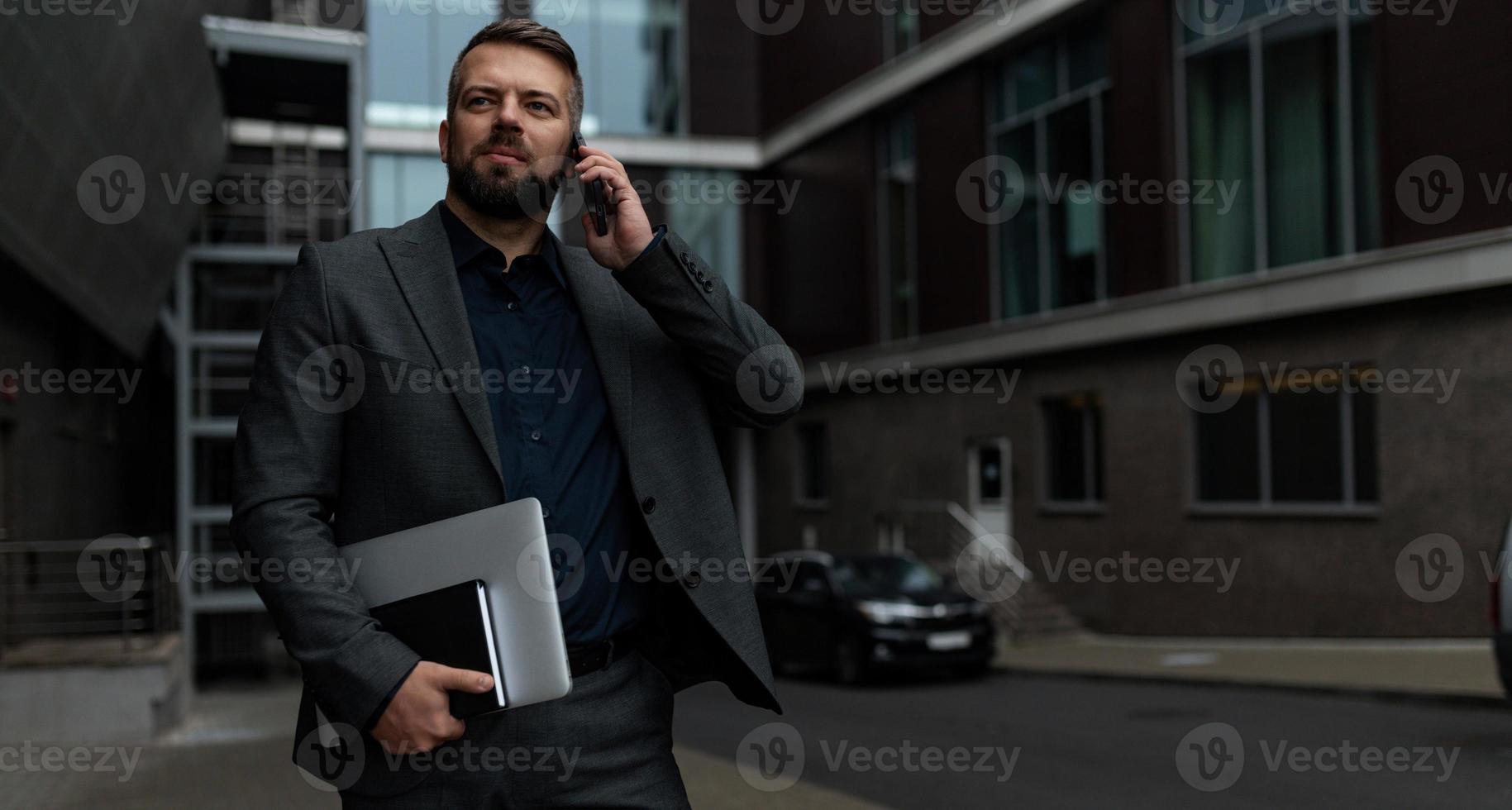 hombre adulto con estilo en un traje de negocios con una computadora portátil en sus manos habla en un teléfono móvil, concepto de una estrategia de marketing exitosa foto