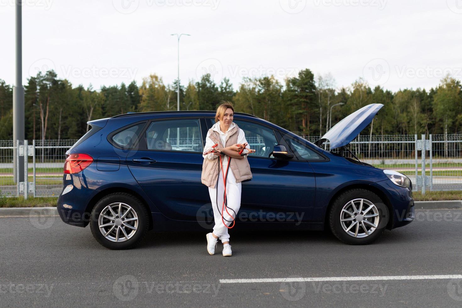 a young woman driver stands in a car with a discharged battery and holds wires in her hands to charge the battery with current photo