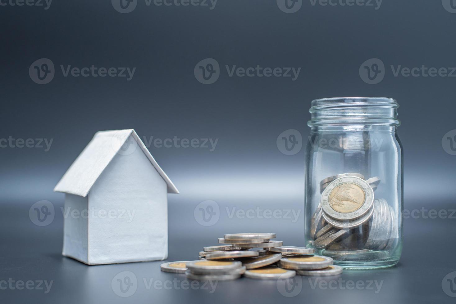 monedas de Tailandia en frasco con monedas de montón sobre fondo negro con modelo de casa foto